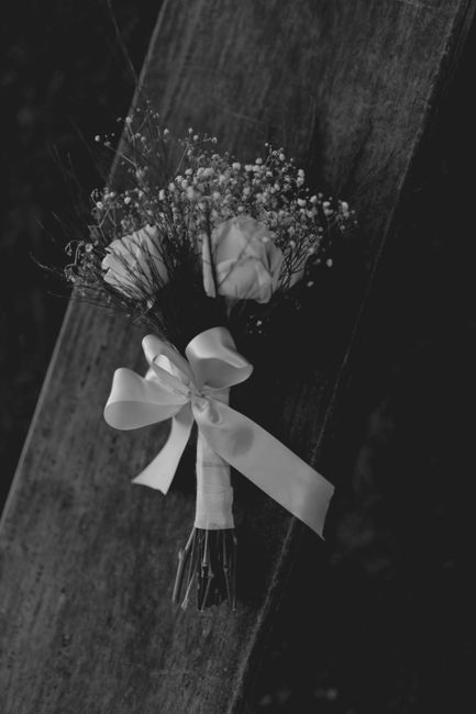 A monochrome photograph of a fresh flower bouquet resting on a wooden surface, featuring roses with delicate petals and small baby's breath flowers with tiny blossoms. The bouquet is wrapped in smooth