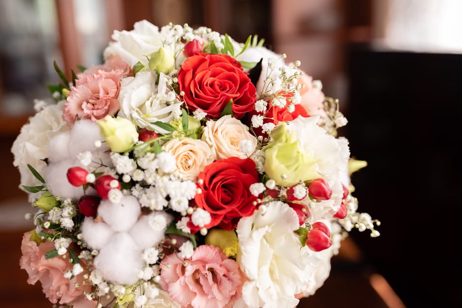 A bouquet arrangement of fresh flowers featuring large red roses, soft peach roses, and white lisianthus, complemented by smaller pink carnations, sprigs of white baby's breath, and green foliage. The