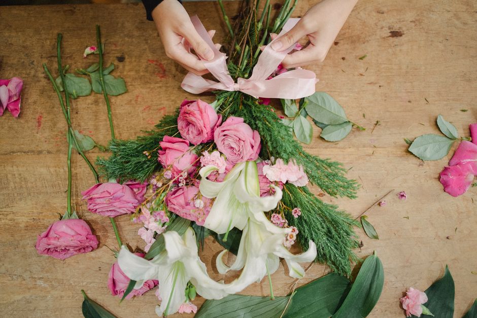A pair of hands arranging a floral bouquet on a wooden table, featuring pink roses, white lilies, and sprigs of greenery, wrapped with soft pink ribbon. Scattered rose petals and leaves surround the b
