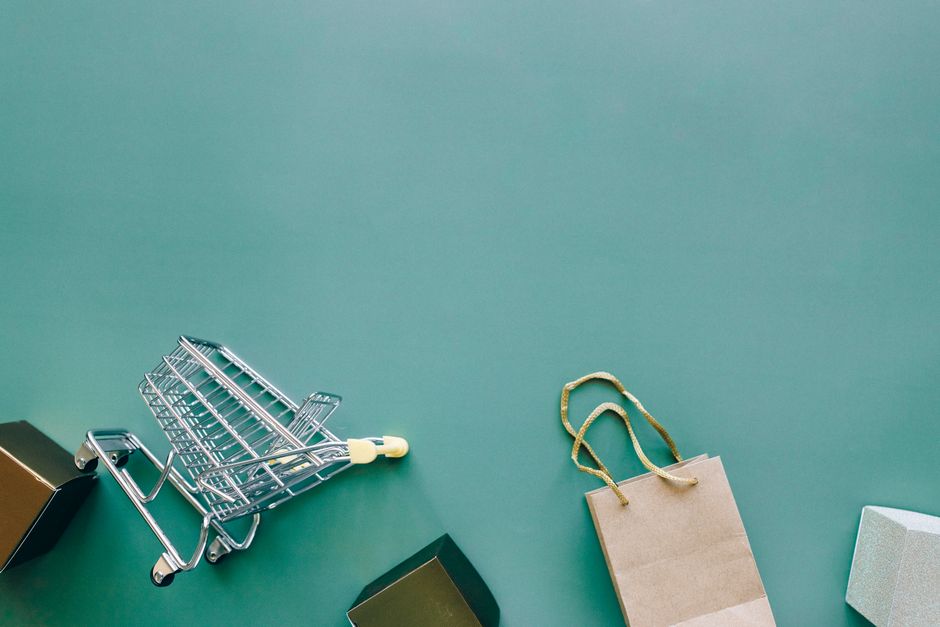 A small, empty metal shopping cart lies on a flat teal surface, with a small beige paper gift bag featuring twisted gold paper handles positioned upright nearby. To the right of the bag, there is a li