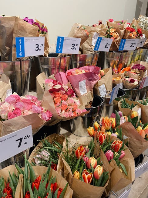 A retail display of various flower bouquets arranged in metal buckets and paper wrapping, featuring a variety of fresh flowers including pink roses, orange and red tulips, and mixed seasonal blooms. E